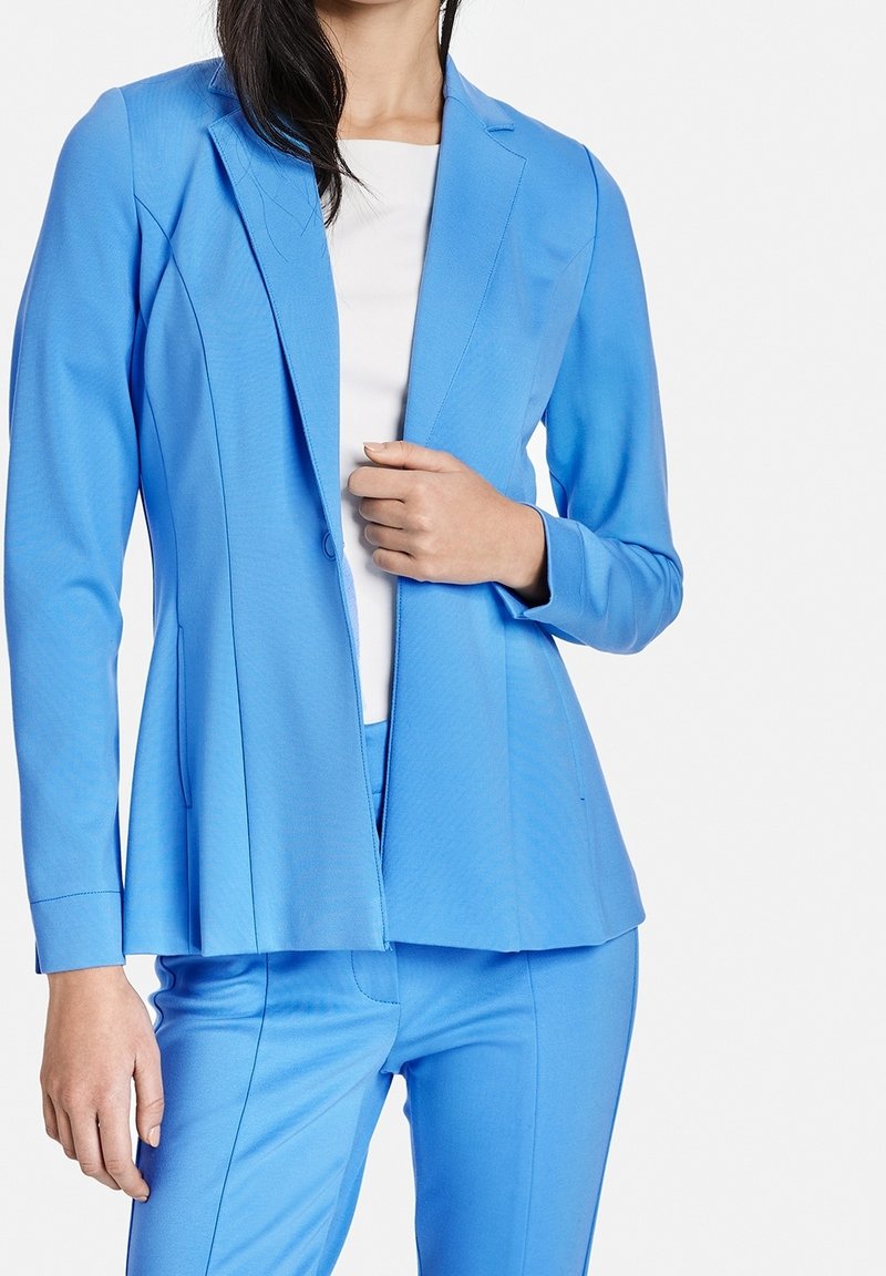 Vaaleansininen räätälöity bleiseri, joka on valmistettu sileästä kankaasta. Siinä on yksittäinen napitus, uurrettu kaulus ja muotoiltu olkapää.