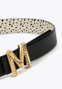 Ceinture en cuir noir avec une boucle en or en forme de "M" torsadée avec des détails texturés ; le côté intérieur présente un fond crème parsemé de taches noires.