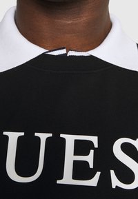 Suéter negro con cuello blanco, con texto en bold en blanco. La tela parece suave, con un detalle en el escote en la parte posterior.