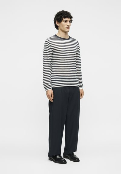 Άνδρας μοντέλο με σγουρά μαλλιά, φοράει μακρυμάνικη μαύρη και λευκή ριγέ μπλούζα, φαρδιά μαύρα παντελόνια και μαύρα επίσημα παπούτσια, στέκεται στραμμένος προς τα δεξιά.