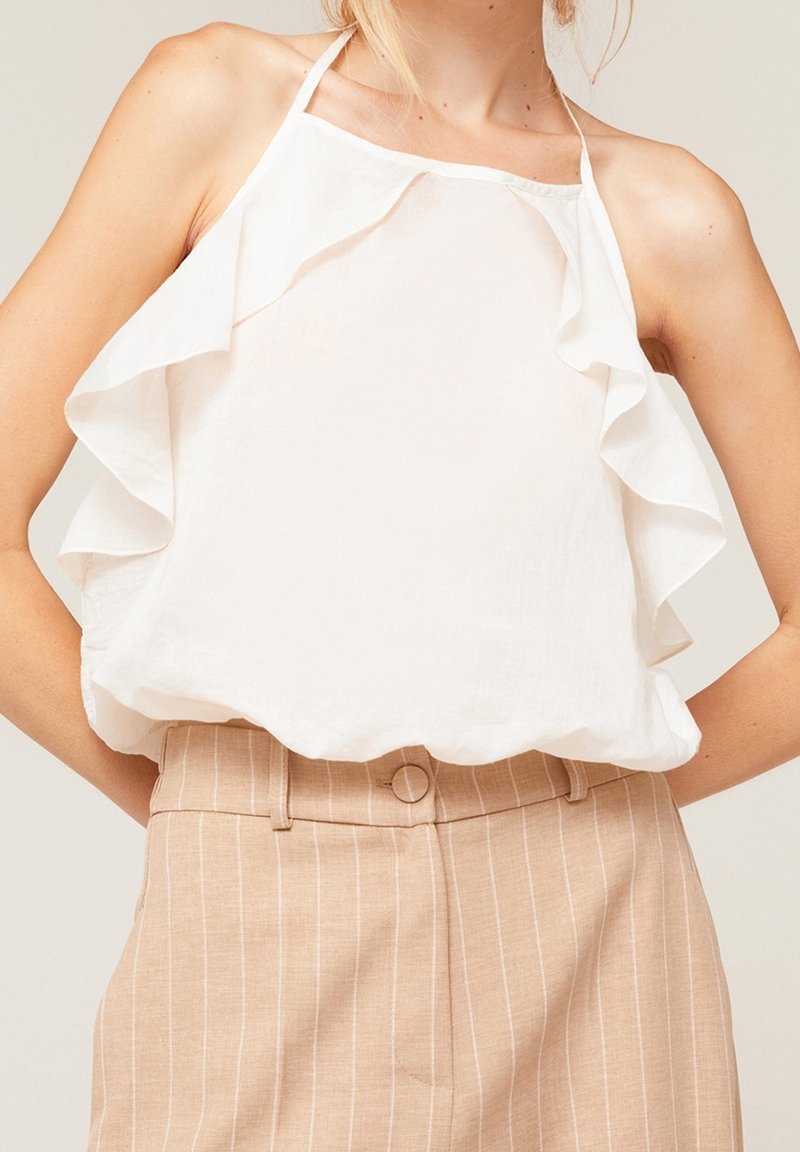 Haut dos nu blanc avec bords froncés, en tissu léger. Associé à un pantalon beige à fines rayures avec une fermeture par bouton.