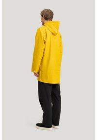 Man i en gul regnjacka med huva, svarta byxor och svarta slip-on skor, stående på sidan framför en ljusgrå bakgrund.