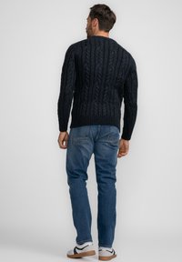 Marinblauer Kabelstrickpullover mit strukturierten Mustern, geripptem Saum, getragen mit blauen Jeans und weißen Sneakers, von hinten betrachtet.