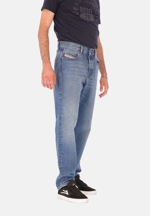 Jean en denim bleu clair avec une coupe droite, doté de cinq poches, de coutures discrètes, et d'une fermeture éclair, porté avec des baskets noires.