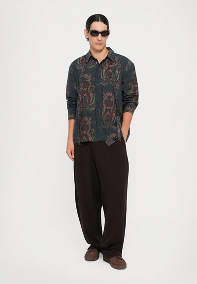 Camicia leggera a fantasia scura con motivi floreali in toni terrosi, abbinata a pantaloni larghi scuri e scarpe marroni.