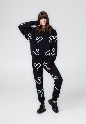 Kobieta w czarnej, dopasowanej bluzie i spodniach z dużymi białymi abstrakcyjnymi wzorami, nosząca czarne trampki, na jednolitym szarym tle.