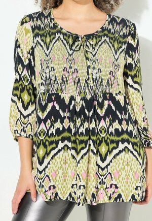 Femme portant une tunique à manches longues avec un motif abstrait vert, noir, blanc et rose et un petit nœud papillon au niveau de l'encolure.
