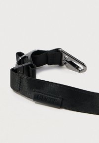 Sangle en nylon noir avec une texture lisse, dotée d'un clip métallique et d'une étiquette logo GUESS en noir embossé.