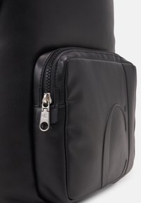 Černý kožený batoh s přední kapsou na zip a stříbrným kováním. Hladký povrch, polostrukturovaný design, s minimálními detaily.