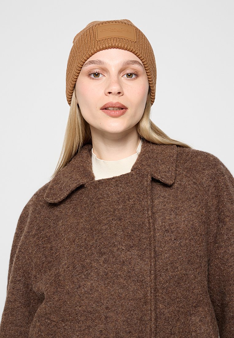 Brun stickad mössa med en texturerad yta och en rektangulär logotyp-lapp. Kombinerad med en löst sittande brun kappa med bred krage.