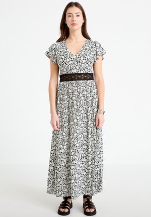 Kvinde iført en sort og hvid blomster maxi-kjole med flagrende ærmer, et sort blonder bælte ved taljen og sorte sandaler med remme, stående stille.