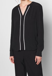 Blusa nera con maniche lunghe, scollatura a V e dettagli in piping verticale bianco. Realizzata in tessuto leggero, presenta una sottile texture e pieghe.
