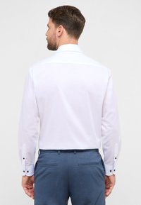 Witte blouse met lange mouwen en een klassieke kraag, voorzien van zwarte knopen aan de manchetten, gedragen met blauwe formele broek. Gladde, lichte stof.