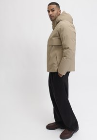 Beige isolierte Jacke mit Kapuze und einer Brusttasche, getragen mit schwarzen weit geschnittenen Hosen und braunen Schuhen vor einem schlichten Hintergrund.