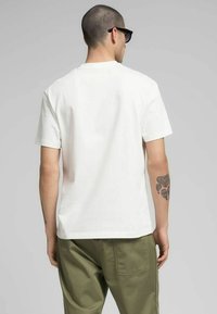 T-shirt bianca a maniche corte con scollo rotondo, in cotone, con vestibilità comoda e tatuaggio visibile sul braccio sinistro. Pantaloni verde oliva.