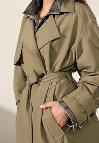 Olijfgroene trenchcoat met een dubbelgebreide ontwerp, grote kraag, ceintuur in de taille en contrasterende donkergrijze binnenkraag en manchetten.