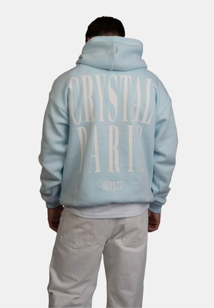 Personne portant un sweat à capuche bleu clair avec un grand texte blanc "CRYSTAL PARIS DEPT. 75" dans le dos, associé à un pantalon de couleur claire.