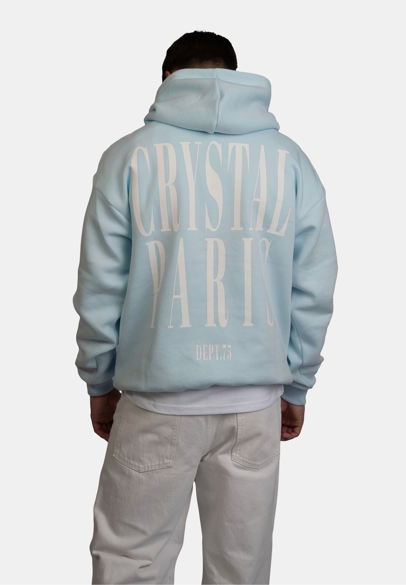 Personne portant un sweat à capuche bleu clair avec un grand texte blanc "CRYSTAL PARIS DEPT. 75" dans le dos, associé à un pantalon de couleur claire.