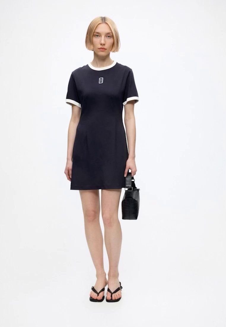 Femme aux cheveux blonds courts portant une robe noire à manches courtes avec bordure blanche, tenant un sac à main noir et portant des sandales noires.