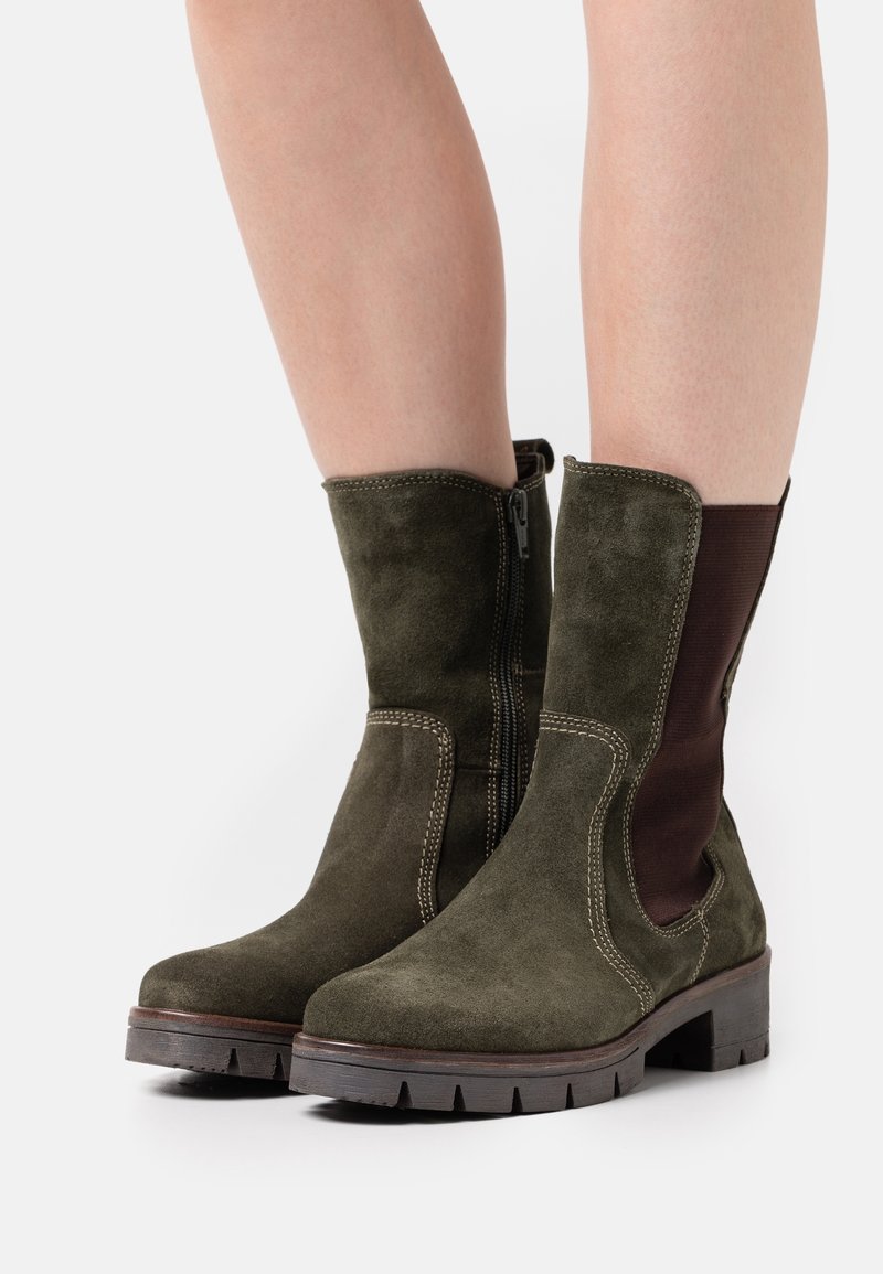 Bottes mi-mollet en suède vert olive foncé avec fermeture éclair latérale, surpiqûres contrastées, panneau arrière élastique et semelle en caoutchouc épaisse, portées sur jambes nues.
