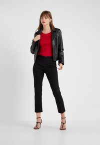 Veste en cuir noire, haut rouge, pantalons noirs ajustés et talons hauts noirs à lanières. Le modèle se tient avec une main sur la veste, sur fond neutre.