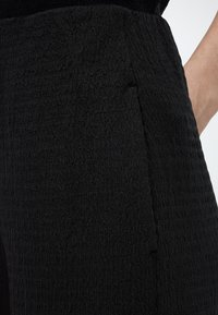 Κοντινό πλάνο ενός ατόμου που φοράει μαύρο παντελόνι με υφή, με ορατή πλαϊνή τσέπη και μερική θέα του χεριού του.