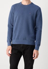 Maglione blu con texture, maniche lunghe e collo a giro. Presenta un piccolo logo sul petto e orli e polsini a costine. Indossato con pantaloni neri.