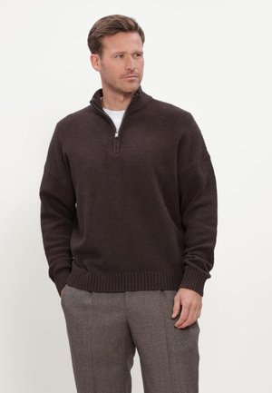 Mann trägt einen dunkelbraunen Pullover mit Viertelreißverschluss und graue Hose, steht mit Händen in den Taschen und schaut seitlich vor einfarbigem Hintergrund.