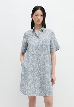 Femme portant une robe-chemise à manches courtes avec un motif floral bleu clair, debout devant un fond blanc uni.
