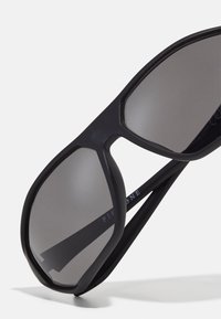Schwarze Sonnenbrille mit einem gewölbten Design, grauen Gläsern und matter Oberfläche. Auffällige Details beinhalten den Markennamen an den Bügeln.