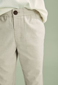 Gros plan sur un pantalon en lin beige avec une taille élastique et un bouton marron, accompagné d'un haut texturé blanc sur un fond vert clair.