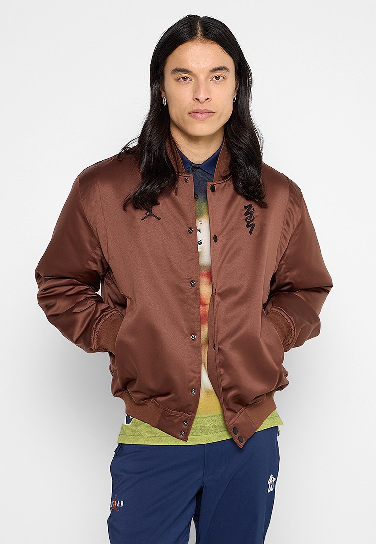 Giacca bomber in satin marrone con chiusura a scatto, polsini e orlo a coste, con patch logo. Abbinata a una camicia grafica colorata e pantaloni blu navy.