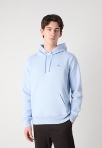 Light blue hoodie made of smooth fabric, featuring a front pocket, drawstrings, and a small logo on the chest. Model paired with dark pants.
