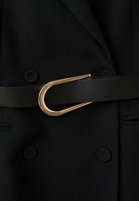Zwarte stoffen kledingstuk met een brede zwarte leren riem, vastgemaakt met een gebogen gouden metalen gesp. Textuurafwerking en twee zichtbare knopen.