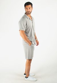 Chemise à boutons à manches courtes gris clair avec une coupe décontractée, assortie à un short. Texture lisse, design subtil, portée avec des baskets blanches.