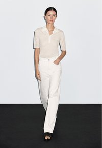 Gebreide polo in lichtcrème met korte mouwen en een knoopsluiting, gecombineerd met witte rechte broek en zwarte sandalen met open tenen.