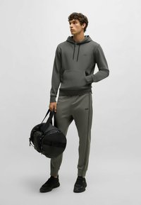 Felpa grigia con cappuccio e pantaloni abbinati con righe laterali, realizzati in tessuto morbido. Il modello tiene una borsa da viaggio nera e indossa sneaker nere.