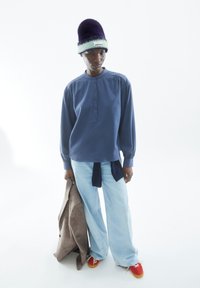 Hellblauses Langarmhemd mit Knopfleiste, kombiniert mit weiter Denim und roten Sneakers. Violette Mütze mit grünem Akzent. Hält eine braune Jacke.