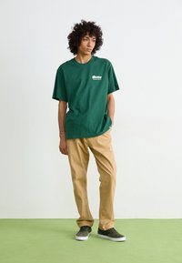 Grøn bomulds-t-shirt med oversized pasform, udstyret med hvidt logo-print. Brune bukser med en afslappet skåret. Grå sneakers med hvide såler.