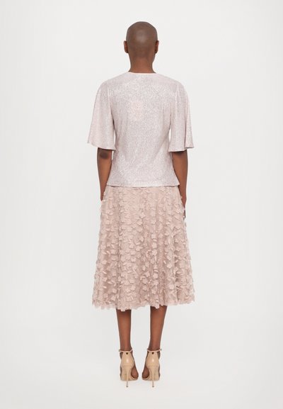 Light pink metallic top with short flutter sleeves, paired with a knee-length skirt featuring floral appliqués. Classic nude heels complement the outfit.
