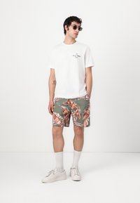 T-shirt en coton blanc avec un petit logo noir, short vert avec un motif de feuilles orange et rouge, baskets blanches et lunettes.