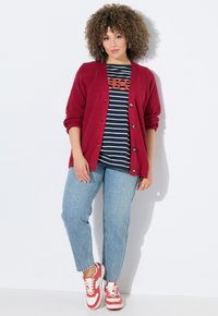 Rot gestreifter Cardigan mit Knöpfen, gestreiftes T-Shirt in Marineblau und Weiß mit rotem Druck "98", hellblaue Jeans und rote Sneakers mit weißen Akzenten.