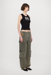 Schwarzes, geripptes Tanktop mit weißem "Moschino Jeans"-Schriftzug, kombiniert mit übergroßen grünen Cargo-Hosen mit Seitentaschen und strukturiertem Stoff.
