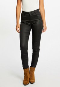 Černé skinny džíny z hladkého materiálu s vysokým pasem, zipovými kapsami a modrým tlačítkem, doplněné béžovými kotníkovými botami.