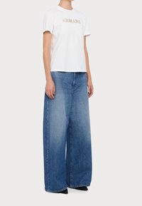 Vit bomullst-shirt med "ARMANI" i guldtext, kombinerad med vida ljusblå jeans och svarta spetsiga skor.