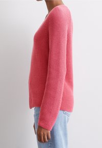 Pull en tricot rose avec un col en V, manches longues et texture côtelée. Coupe légèrement ample, présenté de côté sur un fond uni.