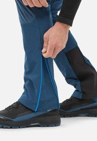 Main en train d'ajuster la fermeture éclair sur la jambe inférieure d'un pantalon imperméable bleu porté avec des chaussures de randonnée noires et bleues.