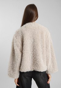 Giacca cropped beige soffice con maniche ampie e un materiale testurizzato simile alla lana morbida. L'orlo è dritto, abbinato a jeans neri.