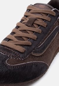 Καφέ αθλητικό παπούτσι Vans με πάνελ από σουέντ και υφή που θυμίζει γούνα, καφέ κορδόνια και ορατές ραφές, σε λευκό φόντο.