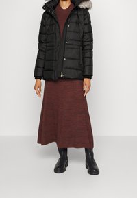 Doudoune noire avec une capuche en fausse fourrure, texture matelassée verticale. Portée par-dessus une robe en tricot bordeaux, assortie à des bottes noires montant jusqu'au genou.